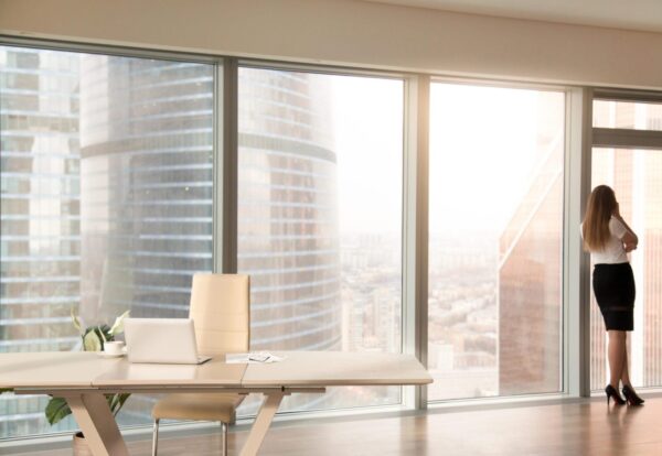 Modern office interior with desk and female silhouette standing back at full length window of business center, talking on phone, looking at cityscape, businesswoman having break at work after meeting